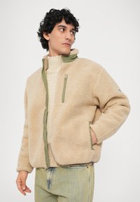 Beige fleece jack met een hoge kraag, groene accenten en een ritszak op de borst. Model draagt een crèmekleurige trui en lichte denim onderkant.