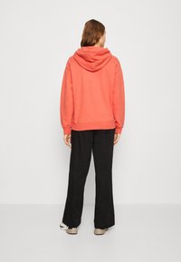 Felpa corallo con cappuccio ampio, vestibilità rilassata, orlo e polsini a coste, abbinata a pantaloni neri a gamba larga e sneakers beige.