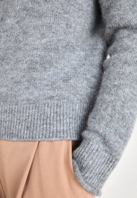 Vaaleanharmaa villapaita, jossa on resorit helmassa ja hihoissa. Se on pehmeä ja hieman väljämallinen. Alla on beige vaatekappale.