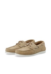 Beige Wildlederbootsschuhe mit weißen Gummisohlen, kontrastierenden Nähten und braunen Schnürsenkeln, die durch Metallösen geführt sind.