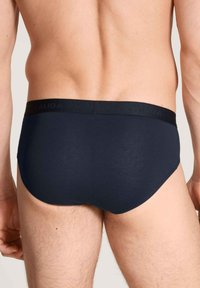 Achteraanzicht van een persoon die een donkerblauwe slip met een zwarte tailleband draagt, tegen een effen lichte achtergrond.