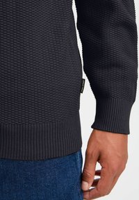 Dunkelblauer, strukturierter Strickpullover mit gerippten Bündchen und Saum. Kleines Markenlabel sichtbar am unteren linken Rand. Nahaufnahme des Ärmels und der Hand.