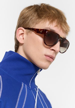 Giovane uomo con capelli castano chiaro che indossa grandi occhiali da sole tartarugati e un top blu a collo alto con dettagli di cuciture bianche.