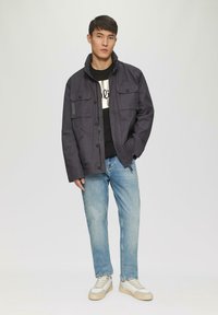 Donkergrijze, kraagjas met vier voorzakken en knoopsluiting, gedragen over een zwart shirt. Lichte spijkerbroek en crèmekleurige sneakers maken de look compleet.