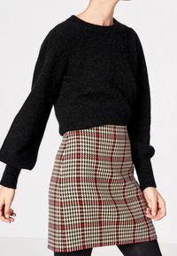 Femme portant un pull noir en peluche à manches longues et une mini-jupe taille haute en pied-de-poule avec un motif à carreaux rouge et beige.