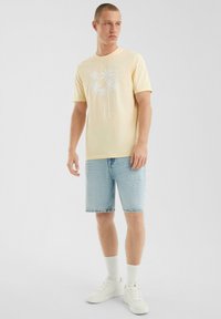Man in een lichtgele t-shirt met palmboomprint, lichte denim shorts, witte crew sokken en witte sneakers, staand op een witte achtergrond.