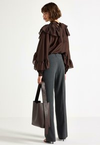 Blusa marrone con maniche a volant, pantaloni grigi sartoriali e una borsa a tracolla in pelle marrone scuro con una tracolla piatta. Tessuto liscio e design strutturato.