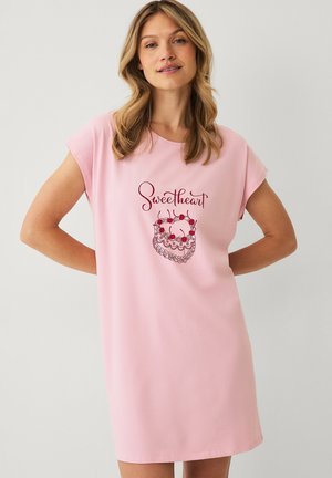 Frau mit blondem Haar trägt ein pinkes Kleid mit dem Text "Sweetheart" und einer herzförmigen Kirschtorte auf der Vorderseite vor einfarbigem Hintergrund.