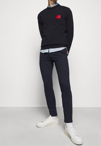 Czarna bluza z czerwoną naszywką logo, jasnoniebieska koszula pod spodem, ciemne spodnie i białe sneakersy. Gładki materiał w luźnym kroju.