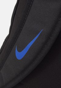 Mochila atlética negra con tejido texturizado, bordes curvados y un destacado logotipo de Nike en azul en el lateral.