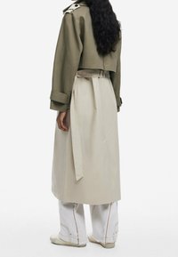 Abrigo trinchera de oliva recortado con una falda beige ceñida a la cintura. Presenta un cuello amplio, detalles de botones y puños ajustados. Pantalones de pierna ancha blancos por debajo.