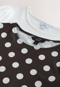 Gros plan sur deux tops de la marque NEXT, un blanc et un noir à gros pois blancs, montrant des encolures rondes et des étiquettes de marque.