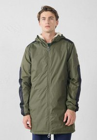 Chaqueta ligera de color verde oliva con mangas negras, cierre de cremallera, capucha ajustable y cuello forrado de forro polar; longitud hasta mitad del muslo.