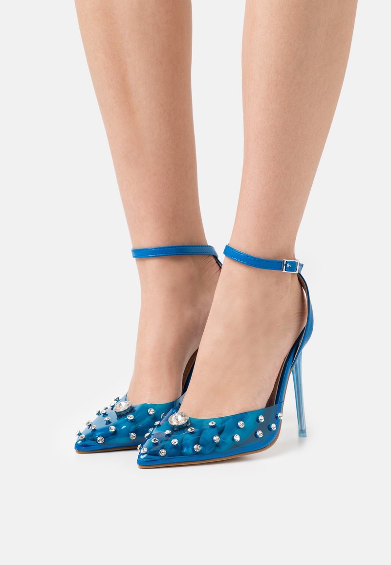 Chaussures bleues à talons hauts avec bout pointu, brides à la cheville et matériau transparent décoré de strass, portées pieds nus sur un fond blanc.