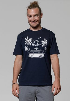 Marineblauwe t-shirt met een witte afbeelding van een vintage bus, palmbomen en tekst: "Laat het avontuur beginnen!" Korte mouwen, ronde hals.