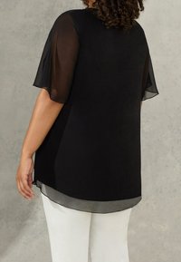 Donna che indossa un blusa nera trasparente a maniche corte con un orlo fluente e pantaloni bianchi, vista di spalle su uno sfondo neutro.