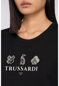 Černé tričko s kulatým výstřihem a výšivkami ruky, dinosaura a štěstěny, se slovem "TRUSSARDI" v bílé barvě.