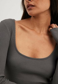 Maglietta grigia a maniche lunghe con costine, dotata di un ampio scollo arrotondato, caratterizzata da una texture liscia e un design aderente, che mette in mostra la pelle nella zona della clavicola.