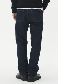 Indigo mėlynos džinsinės kelnės su tiesia pjūvio linija ir matomomis siūlėmis. Yra dvi užpakalinės kišenės ir derinamos su juodais avalynės.