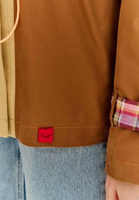 Hnedá bunda s oranžovým prešívaním, s červenou nášivkou s dizajnom rias, kombinovaná so svetlomodrými džínsami a károvanými akcentmi.