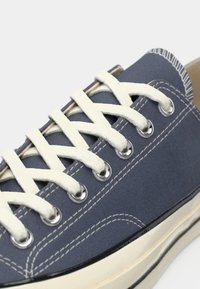Adidași din pânză de culoare albastru închis, cu șireturi albe, ochiuri din metal argintiu și cusături contrastante, având o talpă din cauciuc și un material texturat.