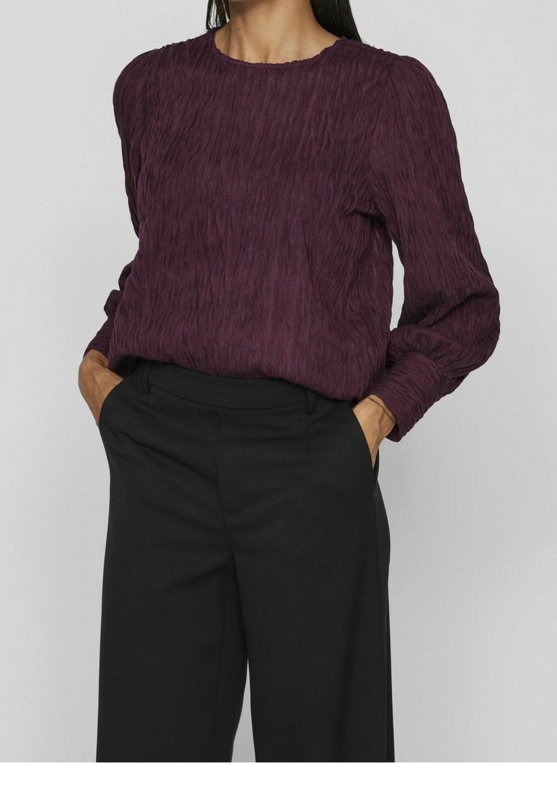 Femme portant un chemisier texturé violet foncé à manches longues, rentré dans un pantalon noir taille haute à jambes larges, les mains dans les poches.