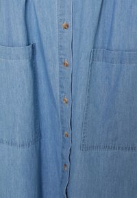 Camicia in denim azzurro chiaro con chiusura frontale a bottoni, due tasche frontali e una texture morbida. Presenta bottoni beige e una vestibilità rilassata.