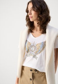 Hvit bomull t-skjorte med metallisk vingsdesign i gull og blått, sammen med en kremfarget strikket cardigan og beige skjørt med knapper.