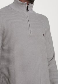 Maglione grigio chiaro con zip, caratterizzato da un motivo tessuto testurizzato. Presenta un colletto alto e un piccolo logo nella zona sinistra del petto.