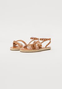 Sandales en cuir brun avec plusieurs bretelles croisées, des clous décoratifs et une semelle en jute naturel. Comprend des brides de cheville réglables.