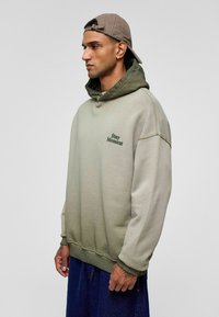 Olivgrüner Kapuzenpullover mit einem helleren Farbverlauf, ausgestattet mit einer Kängurutasche und dunkelgrünem „Stay Moment“-Stick.