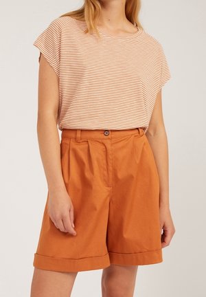 Vrouw met een beige-wit gestreept shirt met korte mouwen, ingestopt in een oranje high-waist short met omgeslagen zomen, staand tegen een effen achtergrond.