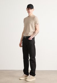 Μπέζ κοντομάνικο μπλουζάκι με διακριτό λογότυπο, μαύρο τζιν με ζώνη, μπέζ παπούτσια και μαύρο καπέλο. Στέκεται μπροστά σε ανοιχτό φόντο.