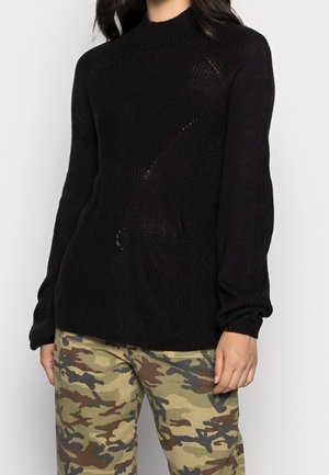 Vrouw draagt een zwarte geribbelde trui met lange mouwen en camouflagebroek, staand tegen een effen lichte achtergrond.