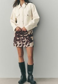 Beskaeret creme denimjakke med to brystlommer, paired med en beige nederdel med et mørkebrun abstrakt mønster og knæhøje sorte støvler.