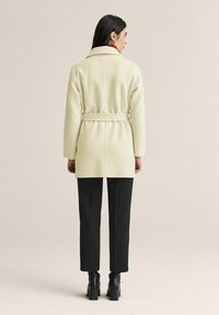 Krämfärgad ullrock med bälte i midjan, slagkrage och långa ärmar. Kvadderad med svarta, raka byxor och klackstövlar.