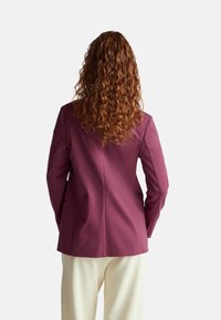 Blazer viola su misura con una silhouette attillata, texture liscia e spalle strutturate, abbinato a pantaloni chiari.