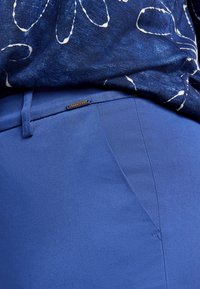 Modré kalhoty s hladkým povrchem, s boční kapsou a kovovým logem. Stylizováno s vzorovaným tmavě modrým topem.