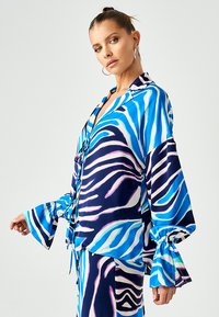 Bluza s plavim i bijelim uzorkom koja ima slobodan kroj, velike zvonaste rukave i kopčanje na gumb, izrađena od glatkog, sjajnog materijala.