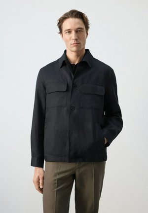 Man wearing a black button-up jacket with chest pockets and brown pants, standing against a plain light background.