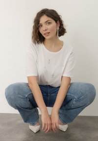 T-shirt bianco con piccoli gioielli decorativi, abbinato a jeans a gamba larga blu chiaro e scarpe argentate a punta. Sfondo semplice.