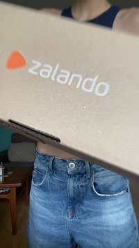 Kartonasta kutija Zalando s bijelim logotipom sa strane. Kutija izgleda malo oštećeno s glatkom teksturom. Tamnoplave traperice su vidljive ispod.
