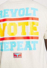 Krémové tričko s výrazným, barevným textem: "REVOLT VOTE REPEAT" v modré, červené a žluté barvě. Obsahuje malé logo na dolním středu.