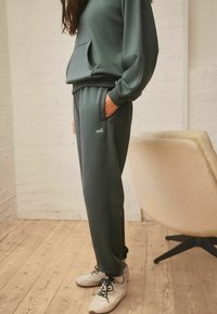 Donkergroen sweatshirt met een voorzak en bijpassende joggers met het woord "self." in witte borduring, gecombineerd met witte sneakers.