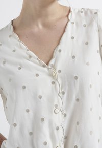 White fabric top featuring a scalloped neckline, silver embroidered dots, and button closures. Soft texture and elegant design details.