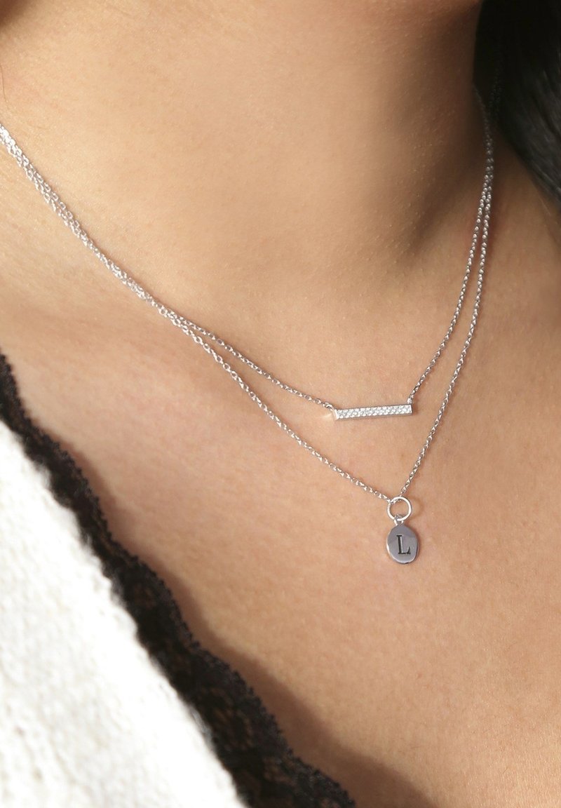 Zilveren dubbele ketting met een horizontale staaf en een ronde hanger met de letter "L," die een delicate kettingontwerp laat zien.