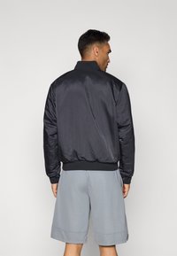 Sort satin bomberjakke med ribbet krave, manchetter og kant. Båret med lysegrå shorts, der viser en glat tekstur og et enkelt design.