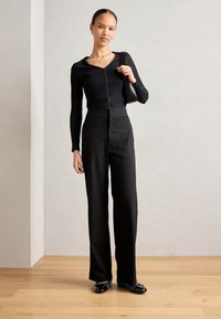 Top nero a coste a maniche lunghe con zip frontale, pantaloni neri a vita alta e ballerine nere in un ambiente interno minimalista con pavimento in legno.