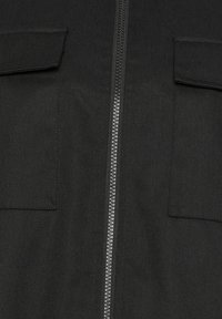 Tissu noir en gros plan montrant une fermeture éclair métallique centrée avec deux poches à rabat symétriques de chaque côté.