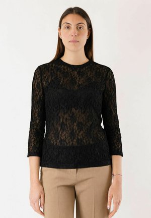 Donna con lunghi capelli castani che indossa una maglia nera trasparente in pizzo a maniche lunghe e pantaloni beige, in piedi contro uno sfondo chiaro e semplice.